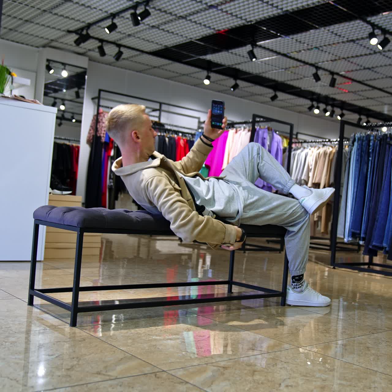 Young man is waiting for his girlfriend near the fitting rooms in the shop. Blonde male lies down on the bench not knowing how to kill time