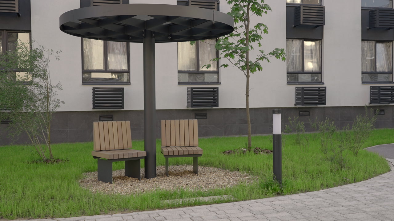 Wooden Seats Under Circular Canopy Shelter, Pebble Bed Island With Young Tree, Paved Pathway And Lamppost