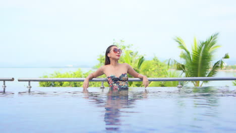 An-attractive-fit-Asian-woman-relaxing-at-a-tropical-resort-in-Thailand,-While-leaning-on-the-edge-of-an-infinity-swimming-pool-slow-motion