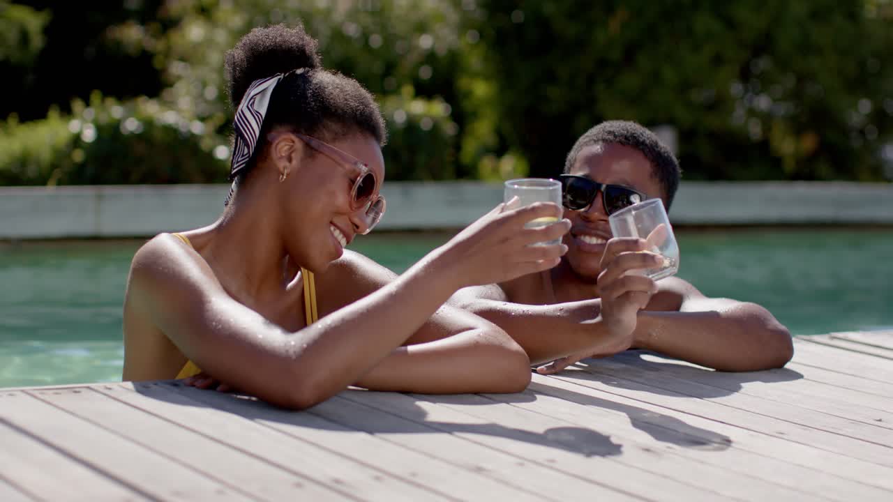 Pareja afroamericana mirándose y girando el uno hacia el otro al borde de la piscina, chocando copas y brindando