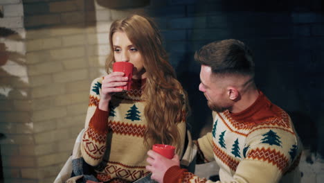 couple in love drinking tea. young man and beautiful girl in knitted christmas sweaters sitting near the fireplace. man kisses his girlfriend. romantic evening.