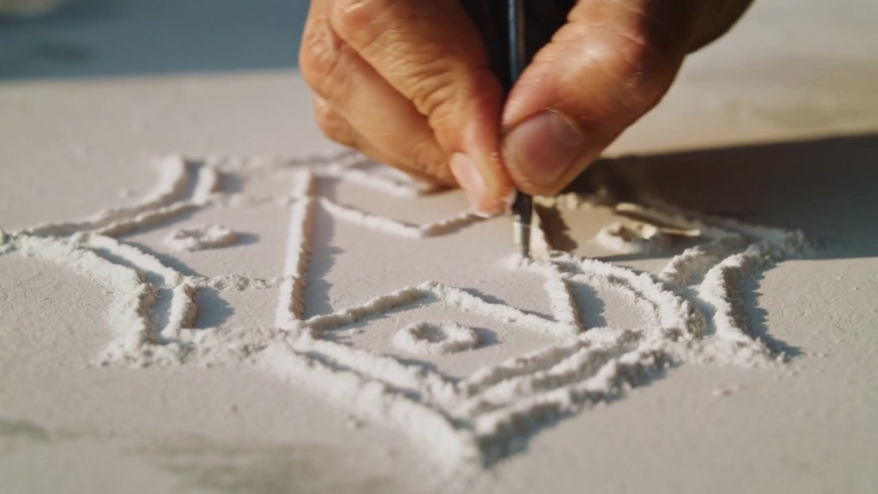 Carving senior hands starting tracing geometric motif in granules on desk, refining edges with tool