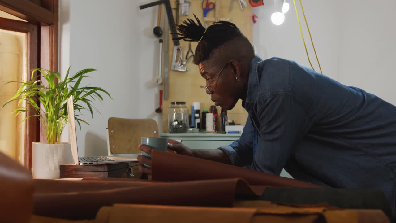 Focused african american craftsman wearing glasses and drinking coffee in leather workshop