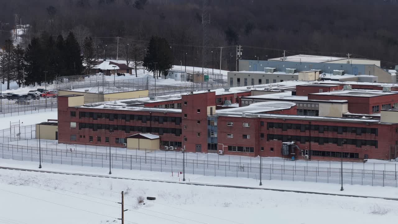 Luftfoto af murstenskomplekset Federal Correctional Institution i Loretto, Pennsylvania, omgivet af sikkerhedshegn og snedækkede områder om vinteren, med skovklædte bakker i baggrunden. Bredt billede.