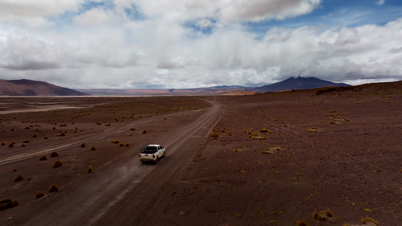 White pickup truck on road trip through vast, remote Siloli Desert, Bolivian Altiplano landscape with mountains, Bolivia. Aerial forward