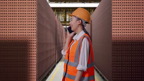 side view of asian female engineer with safety helmet talking on smartphone while standing with red brick packed in stacks are stored