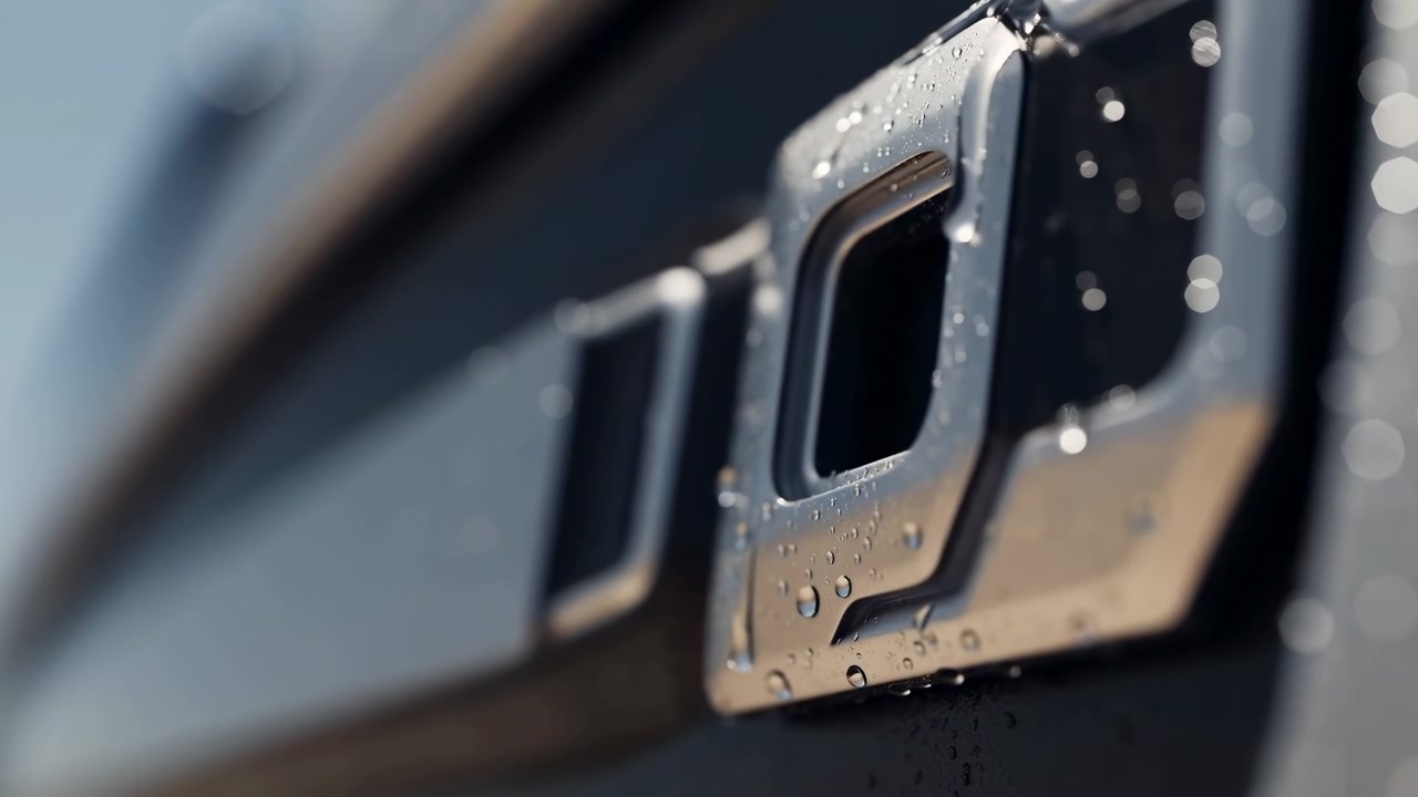 Primo piano estremo di una griglia cromata lucida di un veicolo che mostra un'apertura rettangolare, goccioline d'acqua che si formano lungo il bordo inferiore, goccioline sospese vicino al lato destro e morbidi riflessi bokeh alla luce del giorno