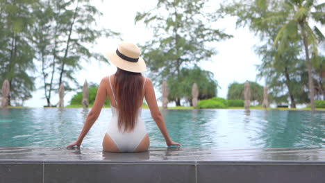 back view of lonely slender young woman at poolside of luxury hotel resort at tropical destination