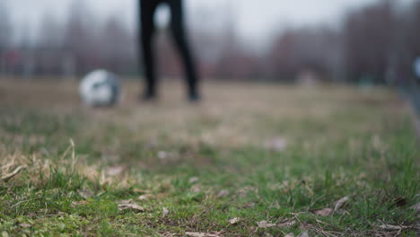 blurred close-up of someone passing a football to another person who traps it under their right foot, the background features a grassy field with a blurred view of people