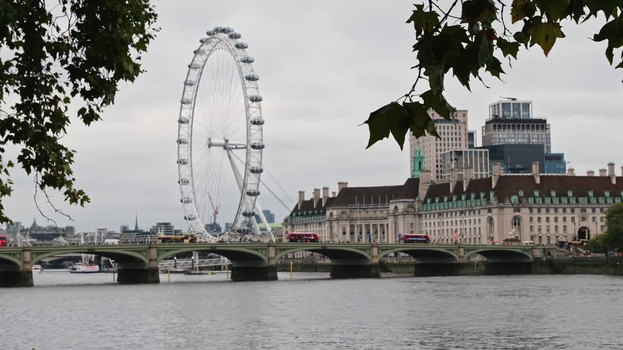 The London Eye observation wheel on the River Thames, stands behind the Westminster bridge as famous red double decker buses drive across, during a cloudy rainy day.