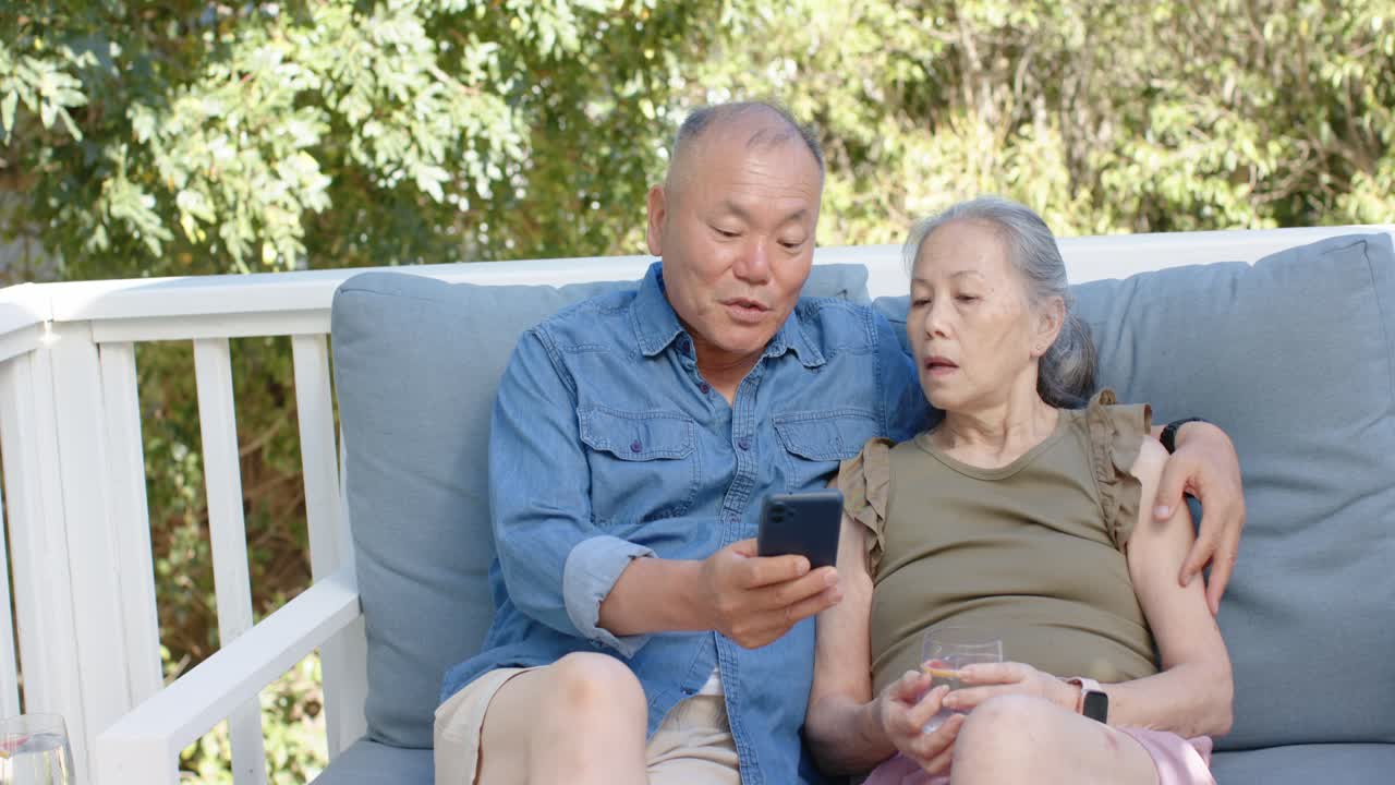 ポーチのベンチに座るアジア人の中年夫婦。男性はスマートフォンを掲げ、コンテンツを共有するためにスクロールしている。