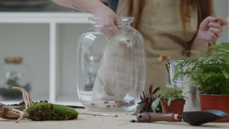 a young woman puts the drainage into a glass jar for creating a tiny live forest ecosystem in it - close-up