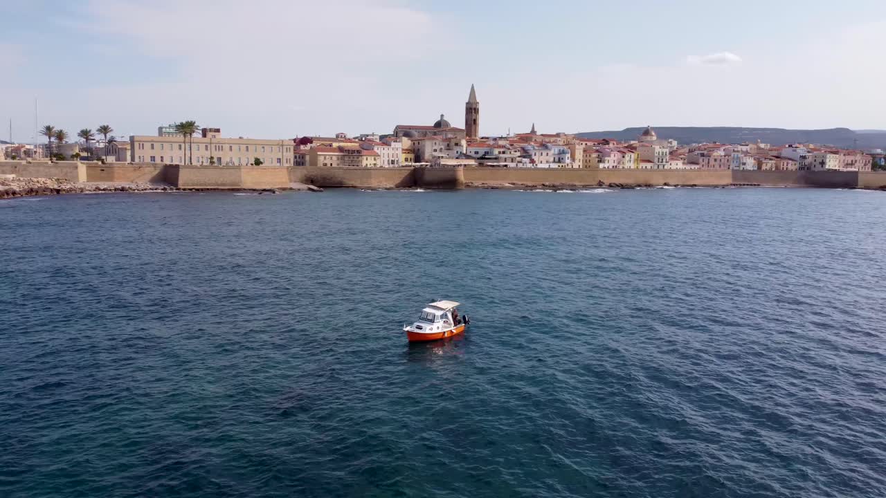 clip de drones sobre el océano con un barco en primer plano y el casco antiguo de alghero, cerdeña en italia