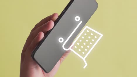 persona que sostiene un teléfono móvil donde aparece la animación de un carrito de compras en línea, fondo amarillo