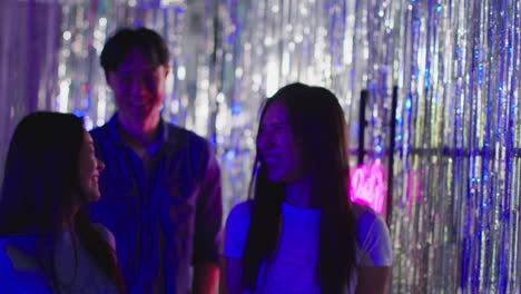 group of young friends walking past neon "let's party" sign hanging against tinsel in nightclub or funfair