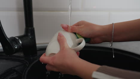 hand view of person wearing wrist chain washing cup with green sponge under running water, cleaning kitchen utensils, hygiene maintenance, household chores, modern kitchen sink