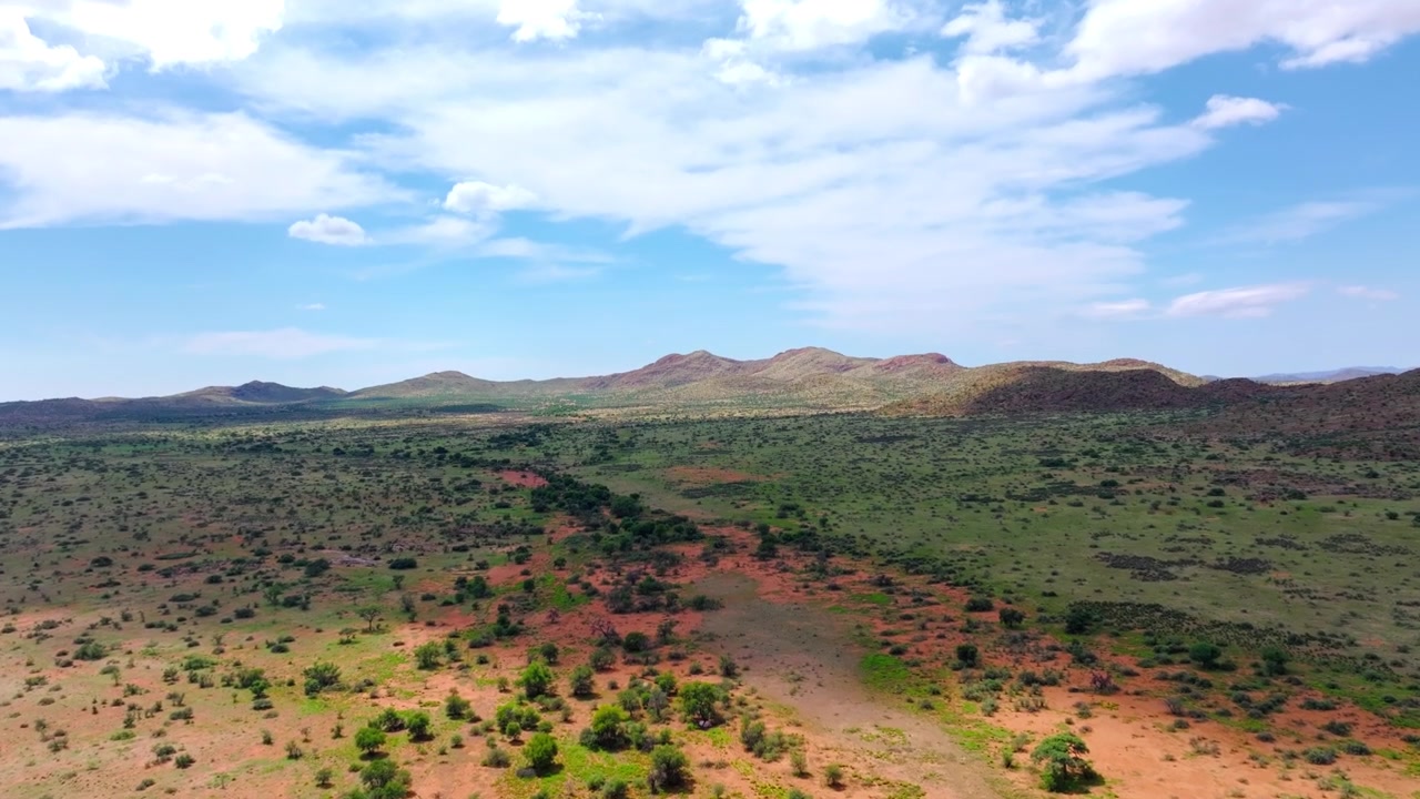 Vista drone su aride colline rocciose e rada boscaglia con terra rossa e una piccola catena montuosa oltre. Colline rocciose brulle e macchia secca si estendono all'orizzonte attraverso questo remoto terreno bruciato dal sole.