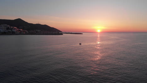 sunrise drone shot with one fishing boat surrounded by serene waves near coastline city with mountain range skyline