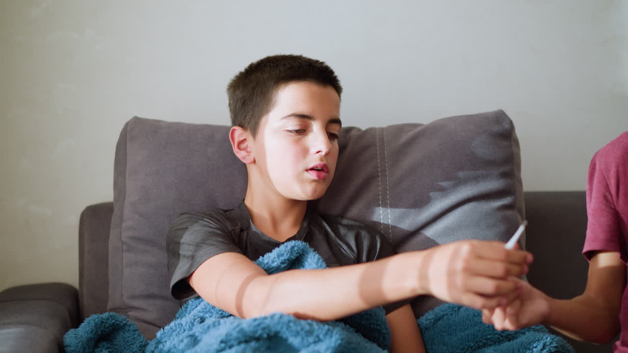 Sick boy sitting on couch reaches out to receive thermometer from sibling to check his temperature while being covered with a cozy blanket, natural light filters through the window