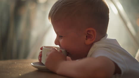 kid smiles playing with cup at table. positive boy leans towards warm drink in room during breakfast. child with ceramic mug sits in sunny kitchen