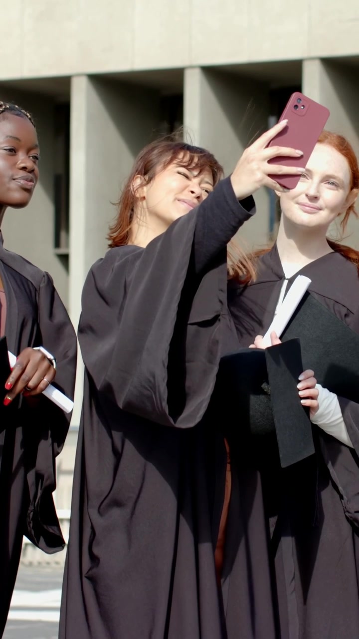 Video verticale: Laureato al centro con toghi e diplomi si fa un selfie con un telefono rosa nel campus.