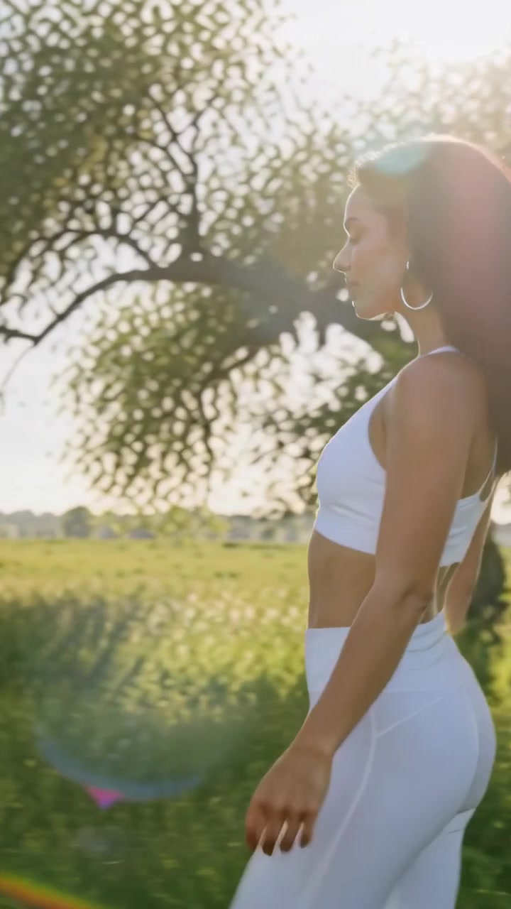 Vídeo vertical: Mulher de leggings brancas caminhando perto de uma árvore no campo, levantando o braço em direção ao sol, com espaço para texto.