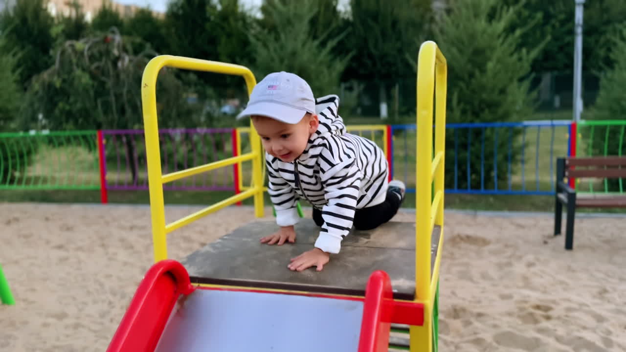 Cute baby boy climbs up to a slide. Cheerful happy Caucasian kid sliding down. Time on the playground.