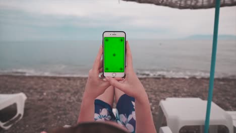 mujer de vacaciones en una cama de sol, cerca del océano y teléfono móvil vertical con pantalla verde, canal alfa y marcas de seguimiento. maquillaje de video de teléfono inteligente y concepto de verano.