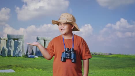 asian boy with a hat and binoculars smiling and pointing to side while traveling in stonehenge. boy researcher examines something, travel tourism adventure, close up