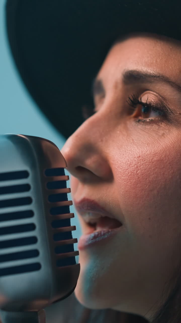 un cantante con un sombrero negro y una chaqueta de cuero negra sostiene un micrófono vintage con una mano mientras hace gestos con la otra, en contra de un fondo azul