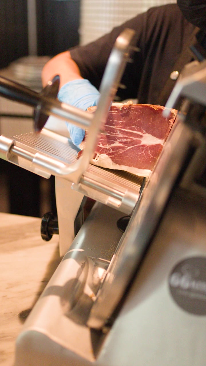 Chef con guanti blu usa un'affettatrice elettrica per fette sottili di prosciutto nella cucina di un ristorante.