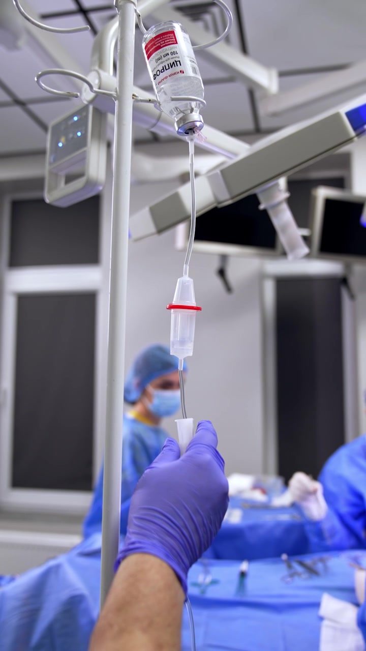 Mani femminili con guanti in lattice regolano il contagocce. Medici in uniforme stanno lavorando al tavolo operatorio sullo sfondo. Video verticale.