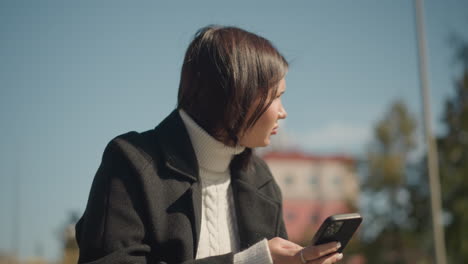 lady seated outdoors, looking thoughtfully while holding phone in hand with ring-adorned finger, blur background features greenery and a building on a bright sunny day