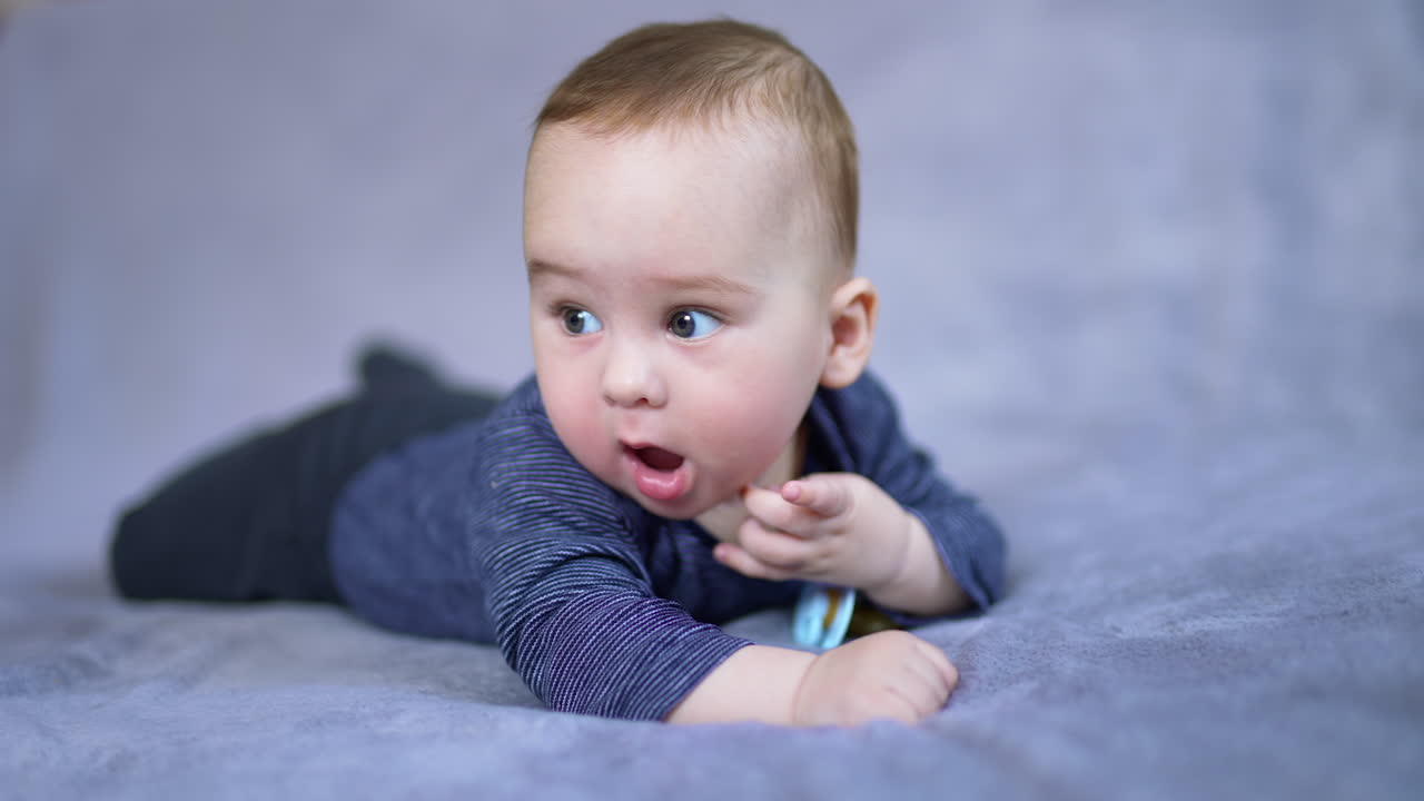 Beautiful baby boy wearing blue shirt lying on his belly. Calm cute toddler shoving tiny plump fingers into mouth. Grey blurred backdrop.