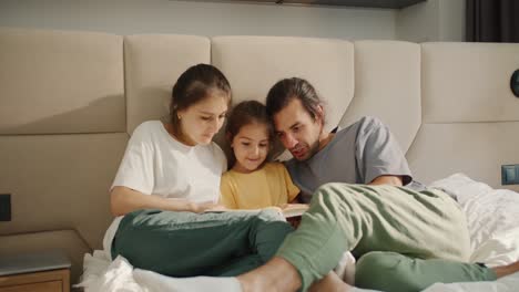 A-little-brunette-girl-in-a-yellow-dress-lies-on-the-sofa-with-her-mom-and-dad-and-reads-a-book-in-a-cozy-modern-room.-A-brunette-man-in-a-gray-T-shirt-and-green-pants,-together-with-his-wife,-a-brunette-girl-in-a-white-T-shirt,-teach-their-little-daughter-to-read-and-looks-at-the-book-Lesha-on-the-