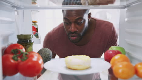 view looking out from inside of refrigerator as man opens door and reaches for unhealthy donut