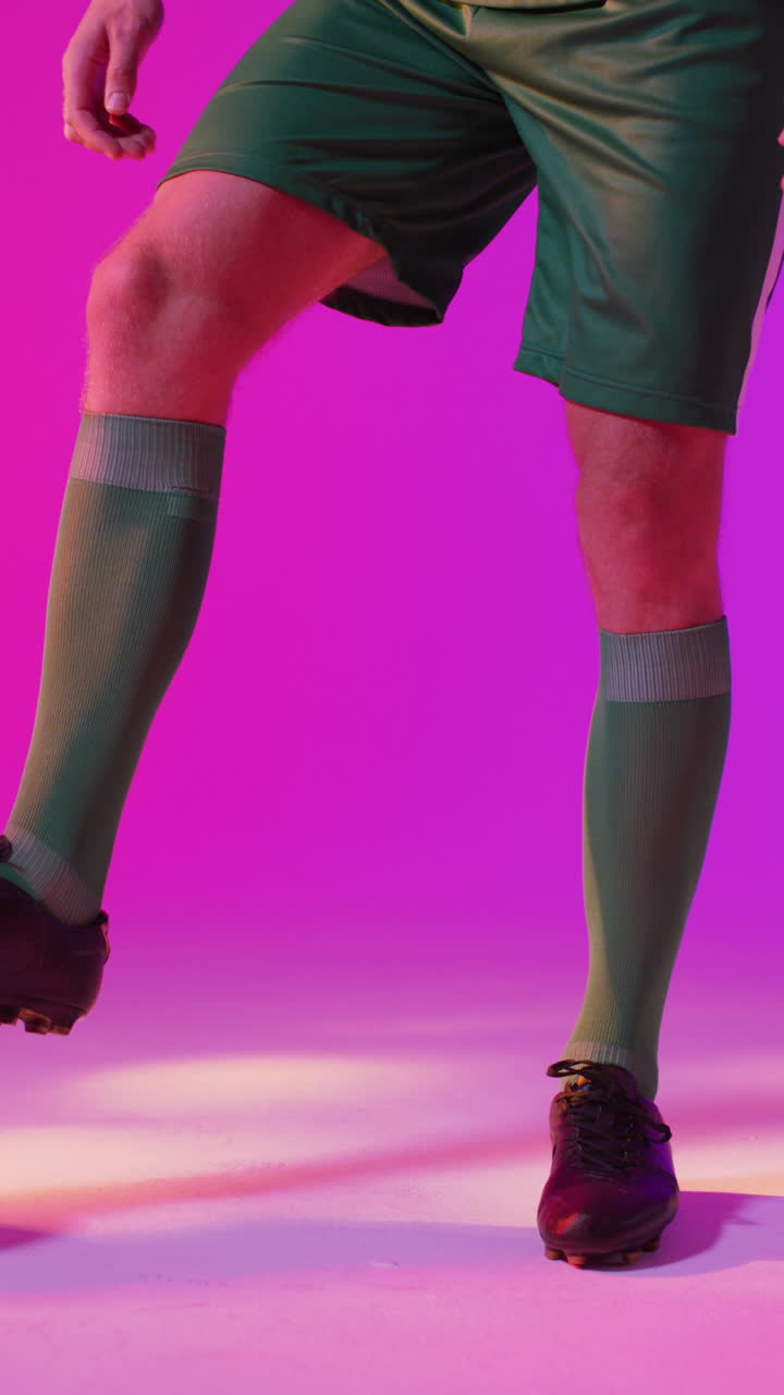 Vertical video of caucasian male soccer player with football over neon pink lighting