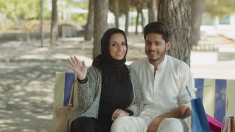 young muslim couple sitting on bench in park, waving to camera.