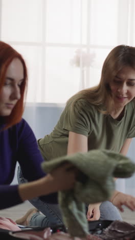 best female friends pack things to go on summer vacation. excited women put clothes into suitcase sitting on floor against bright window slow motion