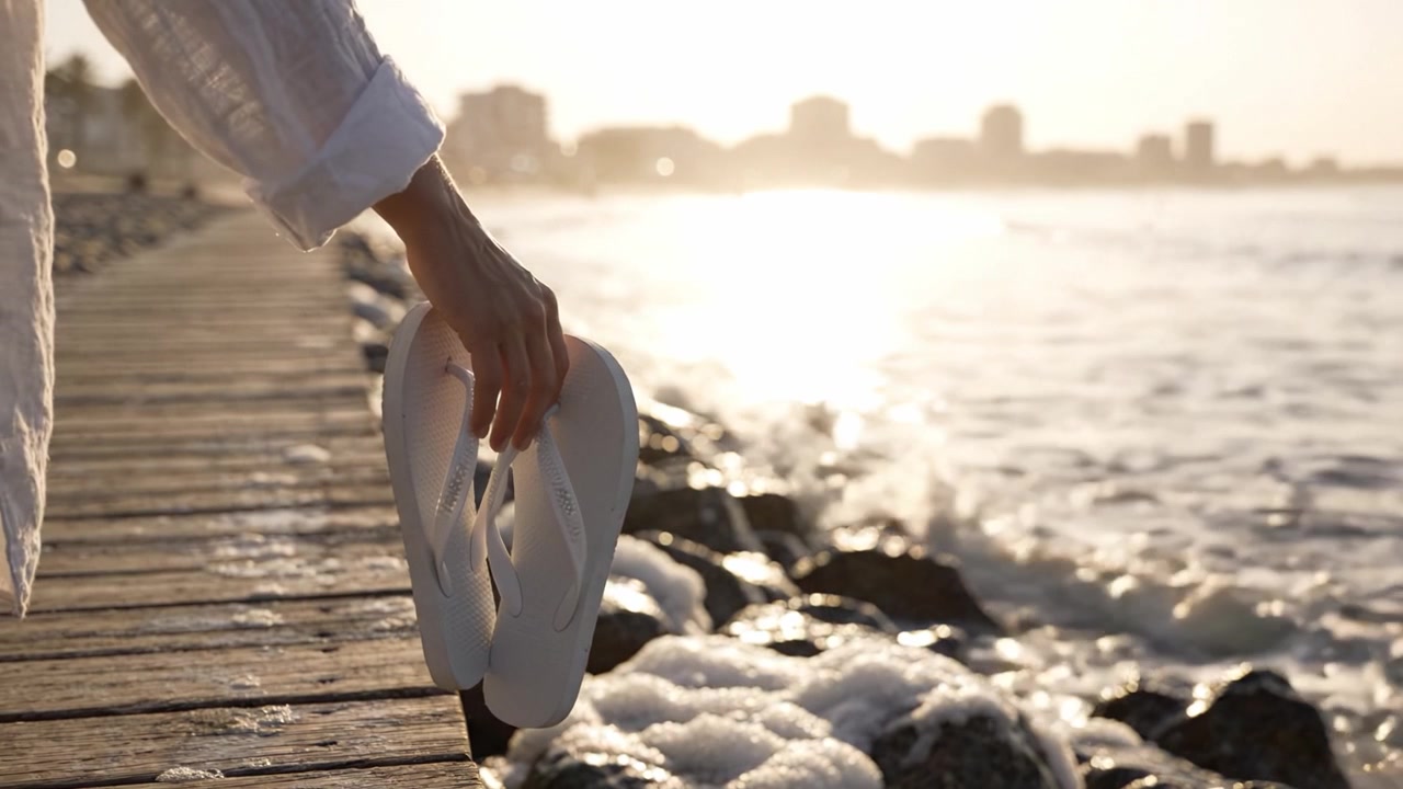 Beatitudine a Piedi Nudi: Un Momento Sereno in Riva al Mare con Infradito Bianche, che Cattura l'Essenza delle Tranquille Serate Estive su una Spiaggia Sabbiosa al Tramonto