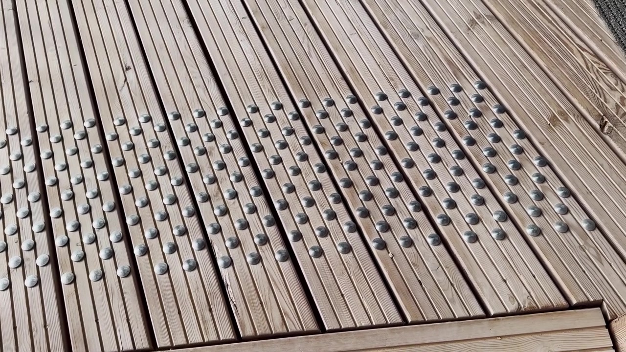 Tactile metal studs on a wooden boardwalk, showing an accessible pedestrian guidance surface with raised warning dots, timber slats, and public pathway design.