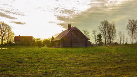 Static-view-of-a-wooden-cottage-with-white-clouds-passing-by-in-timelapse,-surrounded-by-green-grasslands-alongside-a-small-lake-on-a-sunny-day