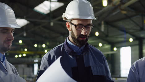 Close-up-view-of-three-engineers-wearing-helmets-holding-blueprint-and-talking-in-a-factory