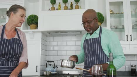 Smiling-senior-diverse-couple-wearing-blue-aprons-and-cooking-in-kitchen