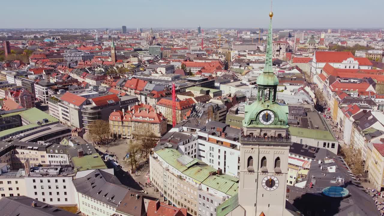 Aerial view of Munich, Germany, with slow drone movement highlighting the tower of Alter Peter and the surrounding urban fabric, revealing red rooftops and historical streets near Marienplatz.