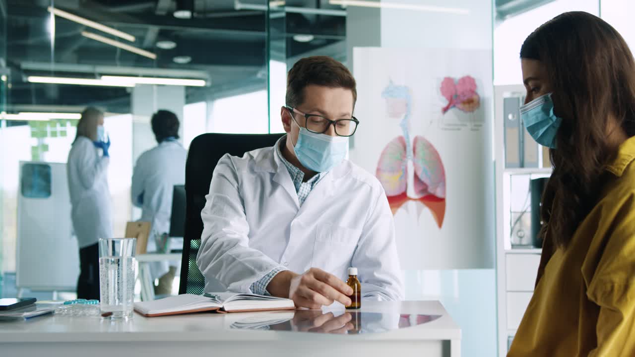 médico caucásico con mascarilla médica sentado en el escritorio y explicando a una paciente el tratamiento del coronavirus en consulta médica