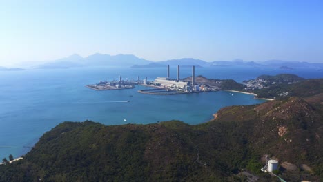 Drone-shot,-traveling-to-the-left,-over-a-big-green-mountain-with-big-coastal-coal-fired-power-plant-on-an-island-next-to-a-village-with-a-beach-during-a-beautiful-sunny-day