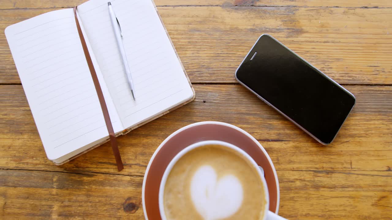 Cup of hot coffee latte with heart shaped foam art with diary and mobile phone 4k