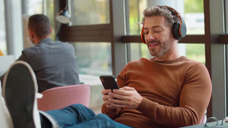 Mature-Businessman-With-Feet-On-Desk-In-Office-Listens-To-Music-On-Wireless-Headphones