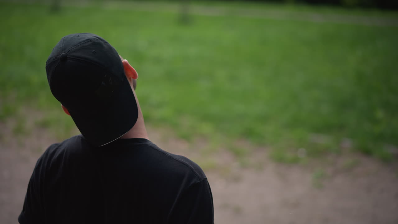 White Man Massaging Neck In Park, Cap Turned Backwards, Rubbing Soreness After Exercise, Calm Green Background, Bench And Trees, Slow Recovery Motion, Focused Breathing And Gentle Stretching